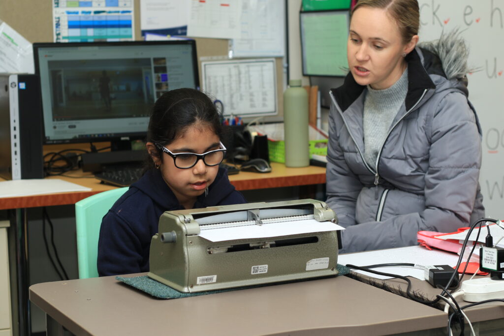 A young girl sitting at a desk typing on a Braille typewriter designed for individuals with vision impairments.